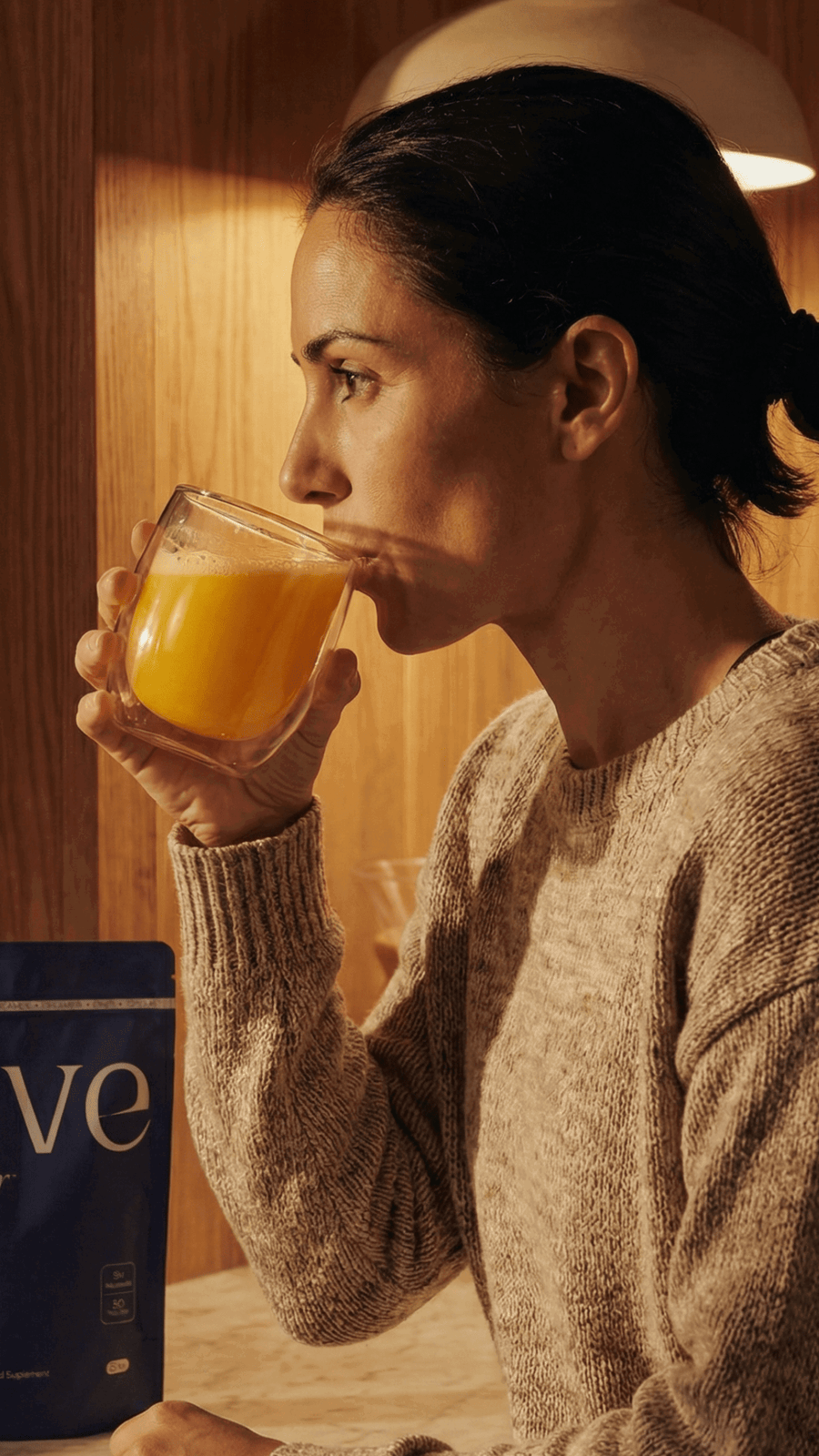 Mujer con jersey de punto bebiendo Dive Dreamer en un vaso de cristal, con la bolsa del producto sobre la mesa